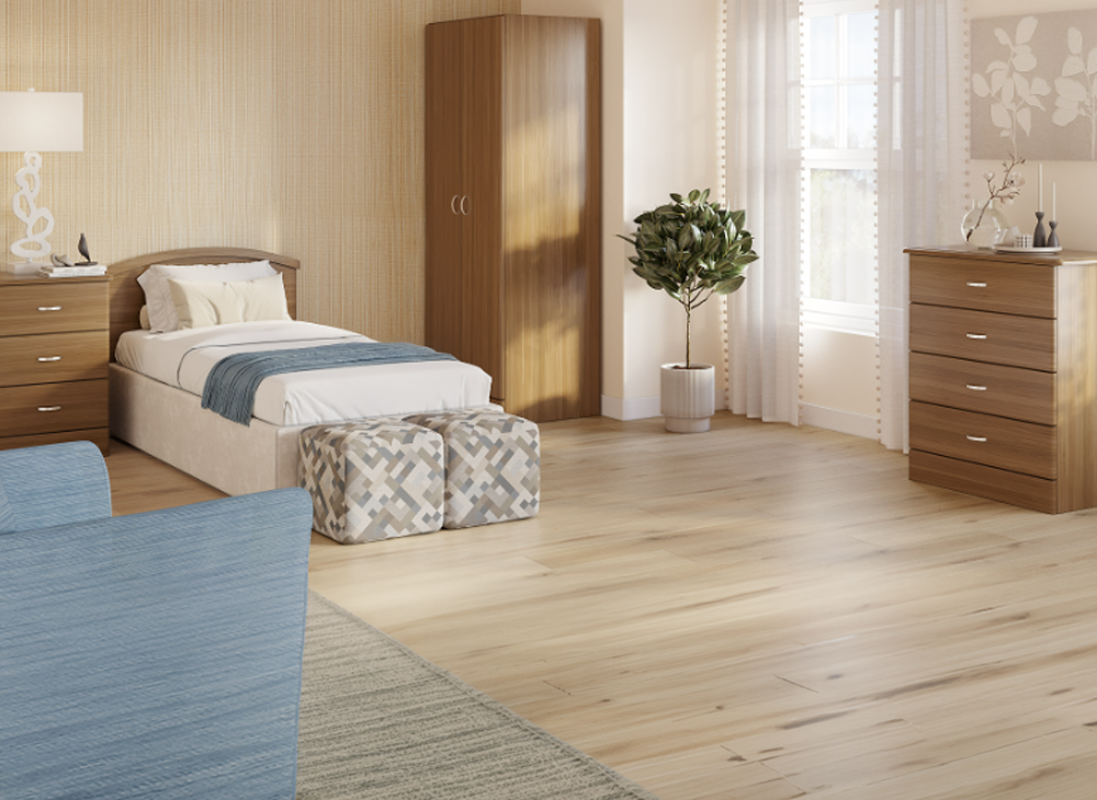 A warm and inviting spacious bedroom that features a colonial blue end chair, a single bed that has a colonial blue blanket to match folded at the bottom of the bed, natural color sheets and pillows. Two cube ottomans that are tan and light blue stand at the foot of the bed. The background has a floor to ceiling rectangular pine wood closet, two dressers: one at hip height with a modern white lamp on top, chest height drawers next to a chest height potted bush that is shaped like a lollypop and light filtering natural floor length curtains.