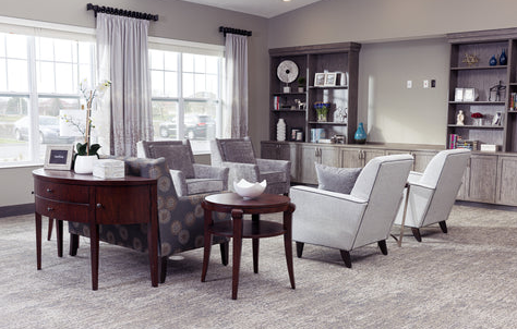 Modern living room with light grey end chairs, sofas both with chestnut wood accents. Beautiful chestnut circular end table in the foreground and matching bookcase cabinet combos in the background.